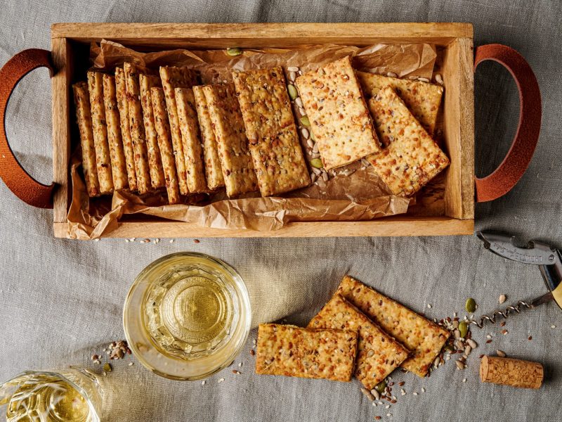 Rustikale Sauerteig-Discard-Cracker mit Körnermix und goldener Kruste neben Weißwein und geöffnetem Korken auf grauem Leinen.