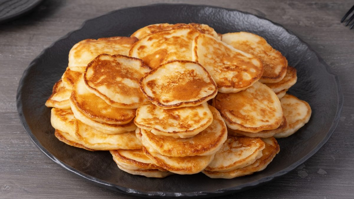 Dicht gestapelte, kleine, goldene Sauerteig-Pfannkuchen mit knusprigen Rändern auf dunkler Tellerfläche vor grauem Hintergrund.
