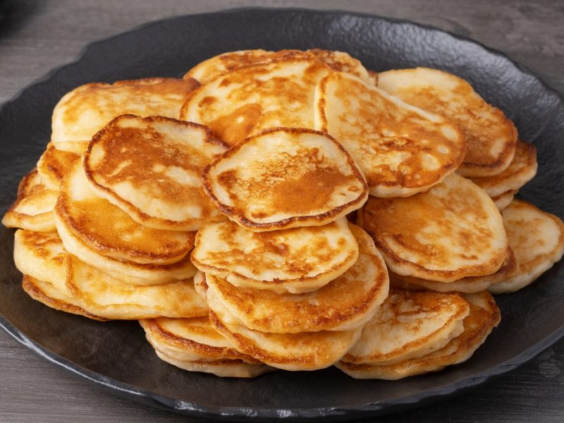 Dicht gestapelte, kleine, goldene Sauerteig-Pfannkuchen mit knusprigen Rändern auf dunkler Tellerfläche vor grauem Hintergrund.