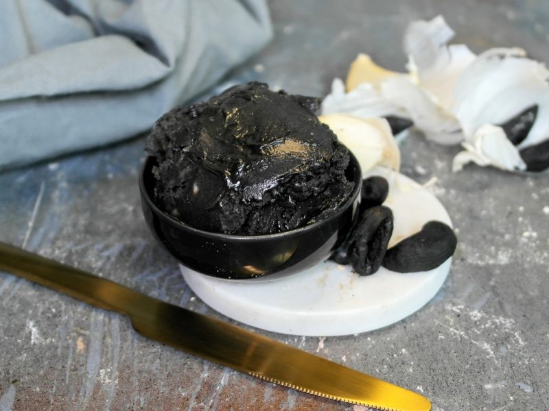 Draufsicht, schräg: Eine kleine Glasschüssel in der ein dunkler Aufstrich ist. Daneben liegen frische Knoblauchzehen.