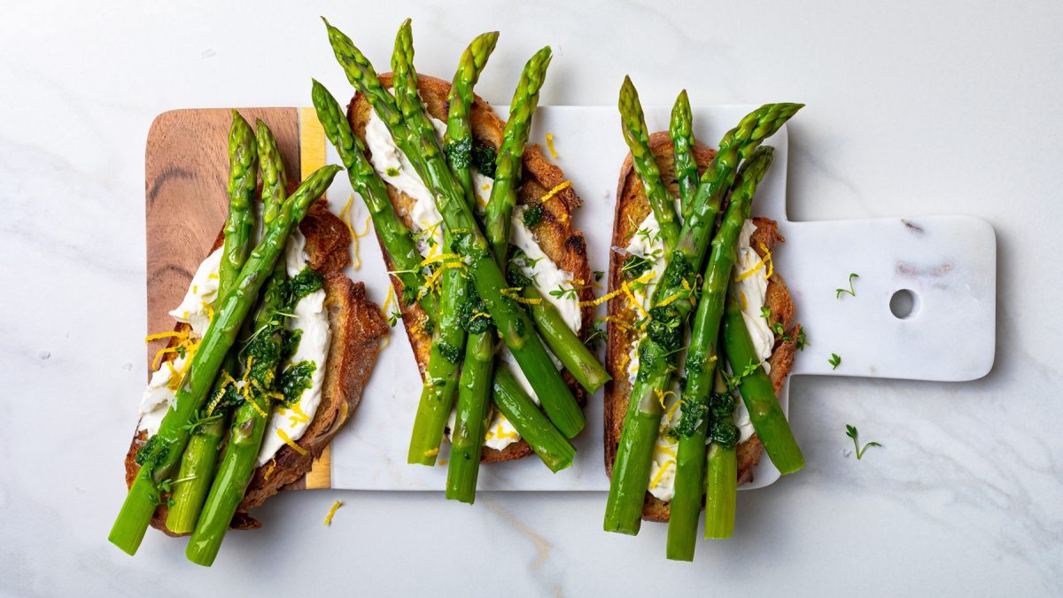 Vier Scheiben geröstetes Brot mit grünem Spargel, cremigem Ricotta, Zitronenzesten und Kräuteröl auf Marmorbrett angerichtet.