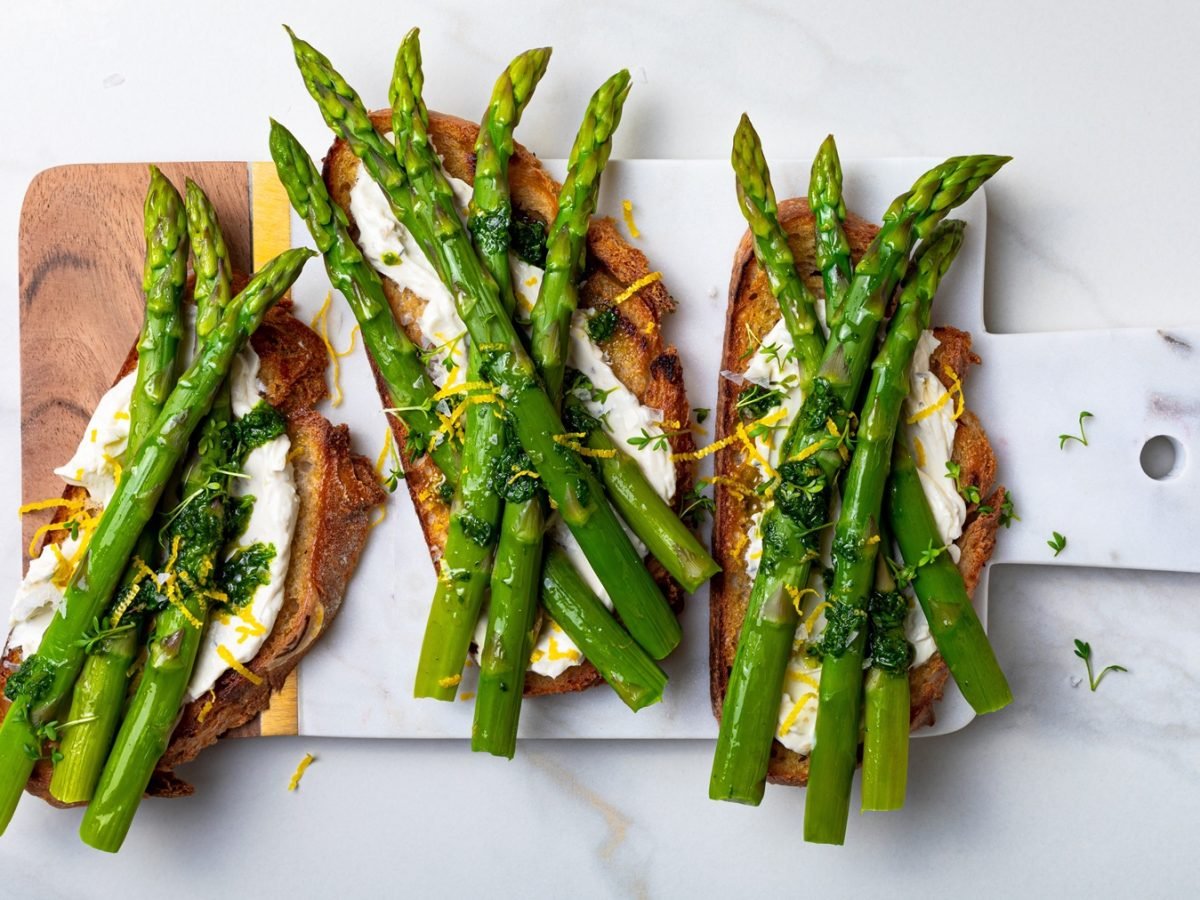 Vier Scheiben geröstetes Brot mit grünem Spargel, cremigem Ricotta, Zitronenzesten und Kräuteröl auf Marmorbrett angerichtet.