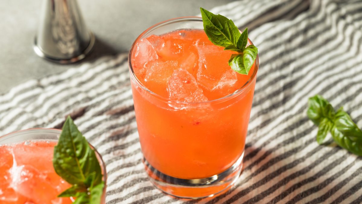 Ein leuchtend roter Strawberry Basil Smash mit Eiswürfeln und frischem Basilikum garniert steht auf einem gestreiften Tuch.