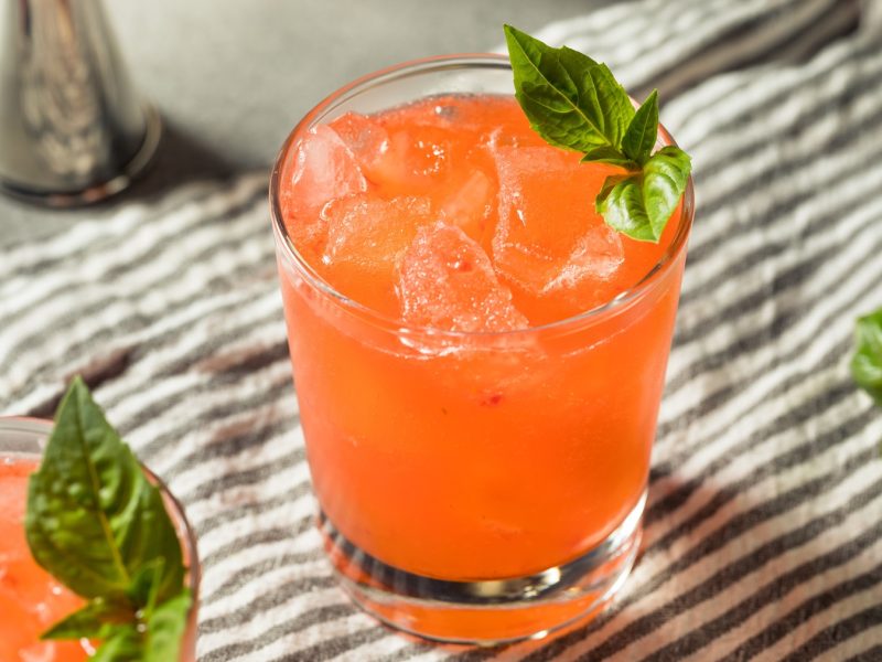 Ein leuchtend roter Strawberry Basil Smash mit Eiswürfeln und frischem Basilikum garniert steht auf einem gestreiften Tuch.