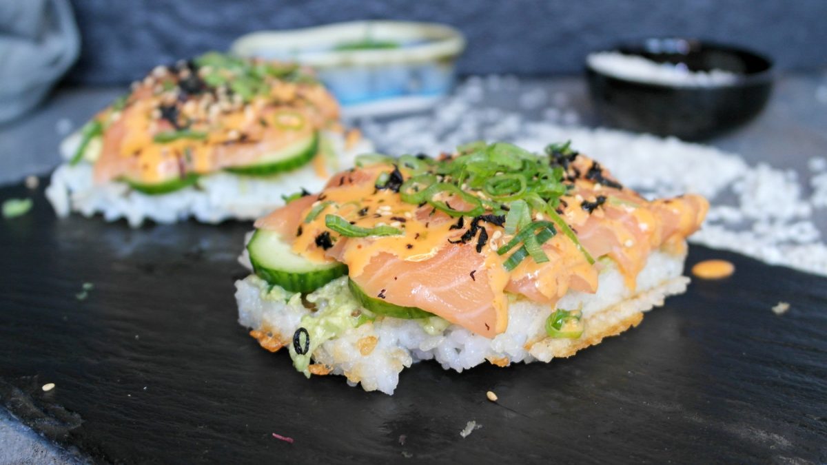 Zwei Reiswaffeln mit Lachs, Avocado und Gurke belegt auf einer schwarzen Schiefertafel und grauem Untergrund.