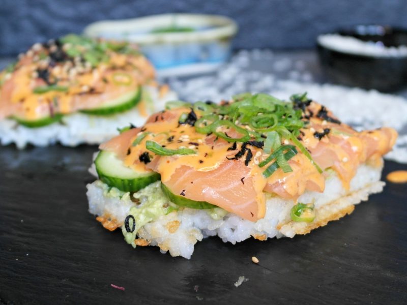 Zwei Reiswaffeln mit Lachs, Avocado und Gurke belegt auf einer schwarzen Schiefertafel und grauem Untergrund.