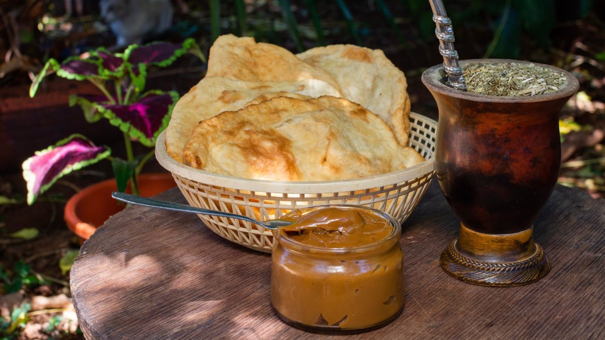 Rustikales Frühstück mit Torta frita, cremiger Dulce de leche und dampfendem Mate unter schattigen Gartenpflanzen.
