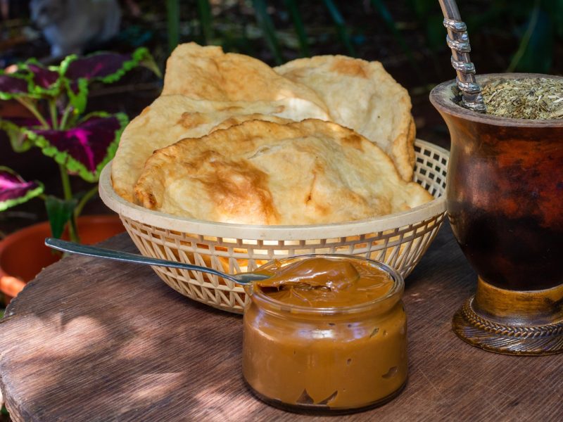 Rustikales Frühstück mit Torta frita, cremiger Dulce de leche und dampfendem Mate unter schattigen Gartenpflanzen.