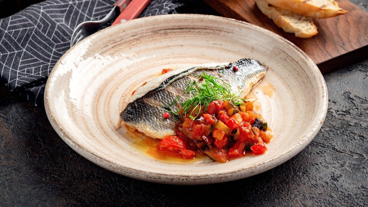 Ein beiger Teller mit dem Gemüse und Fisch auf einem schwarzen Tisch mit Tischdecke im Hintergrund.