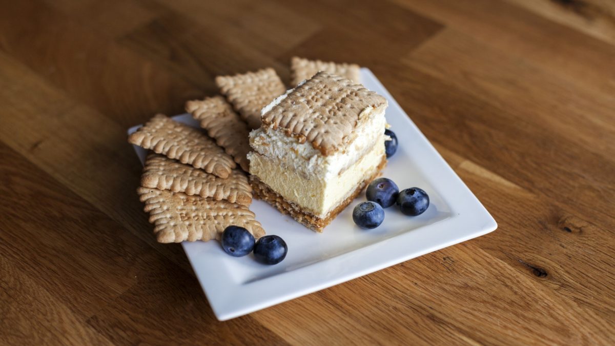 Ein Stück polnischer 3-Bit-Kuchen mit Keksboden, Sahneschicht und Butterkeksen, serviert mit frischen Blaubeeren auf weißem Teller.