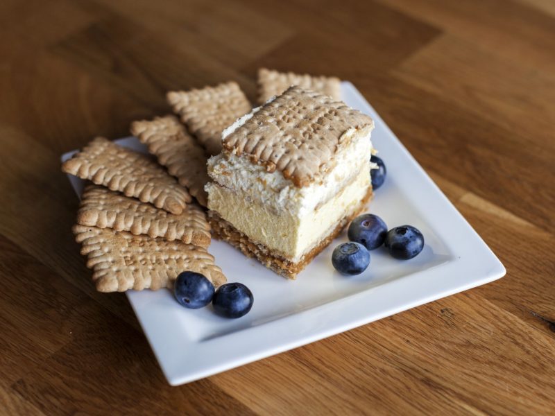 Ein Stück polnischer 3-Bit-Kuchen mit Keksboden, Sahneschicht und Butterkeksen, serviert mit frischen Blaubeeren auf weißem Teller.