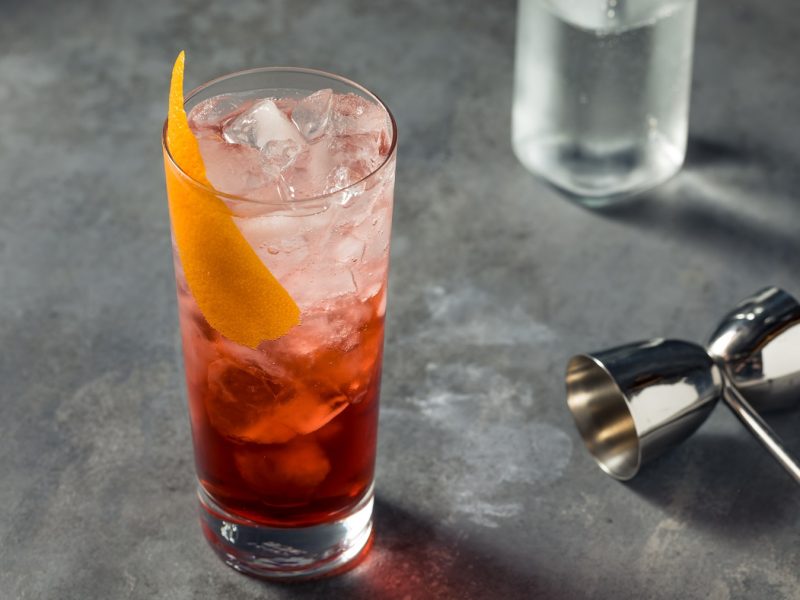 Ein Glas mit dem rötlichen Cocktail auf einem grauen Tisch mit Bar-Equipment von schräg oben fotografiert.
