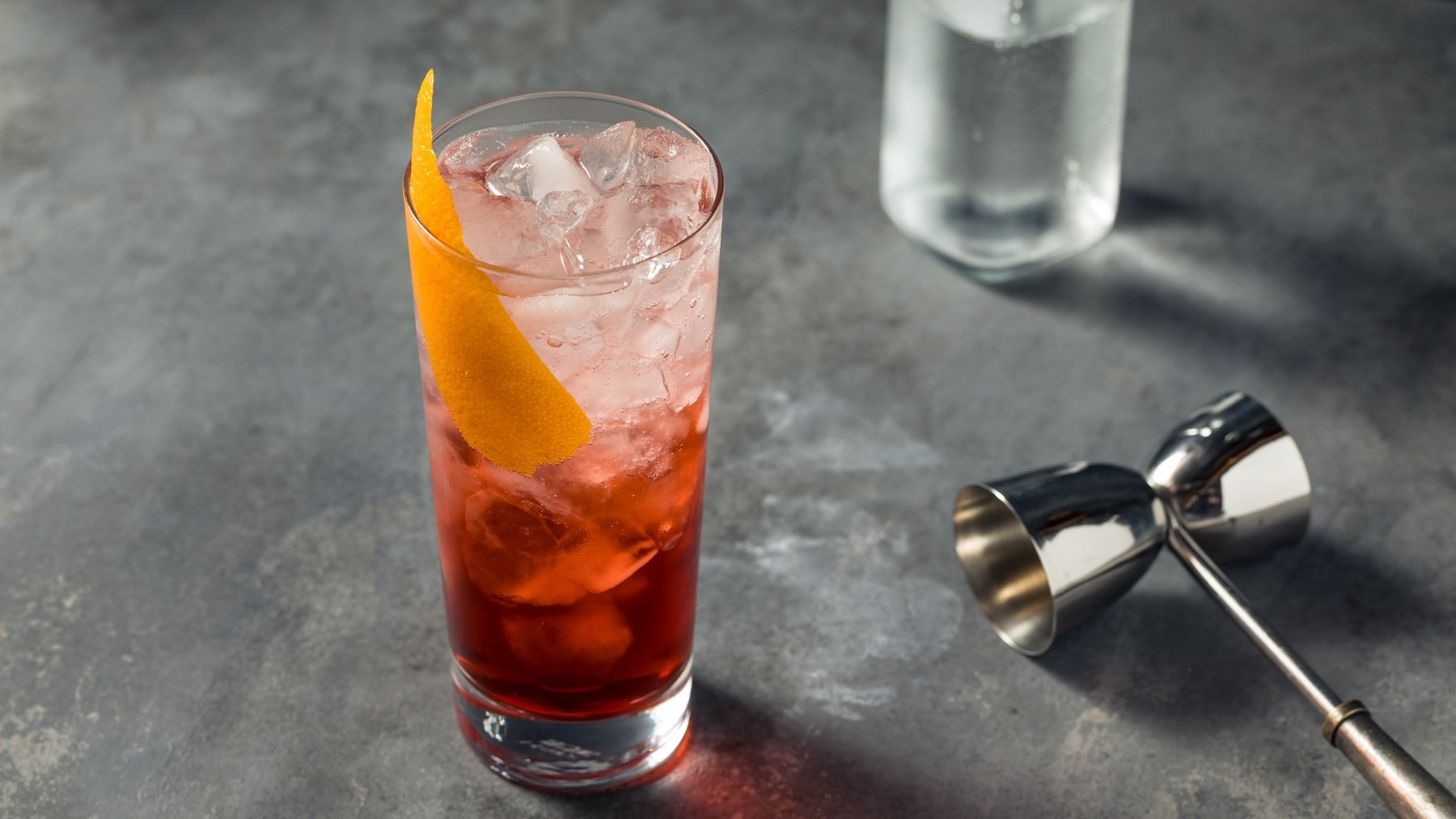 Ein Glas mit dem rötlichen Cocktail auf einem grauen Tisch mit Bar-Equipment von schräg oben fotografiert.