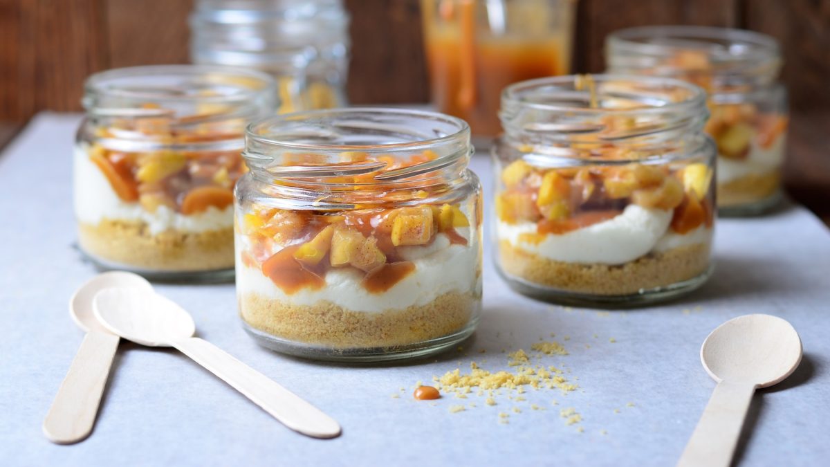 Vier Gläser mit dem Trifle auf einem hellblauen Tisch mit einer Holzwand im Hintergrund. Alles von der Seite fotografiert.