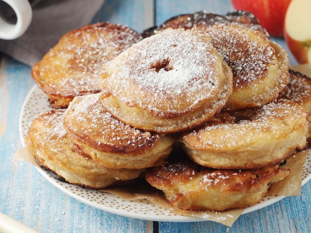 Mehrere Apfelplinsen mit Puderzucker bestäubt auf einem gemusterten Teller, daneben ein angeschnittener Apfel.
