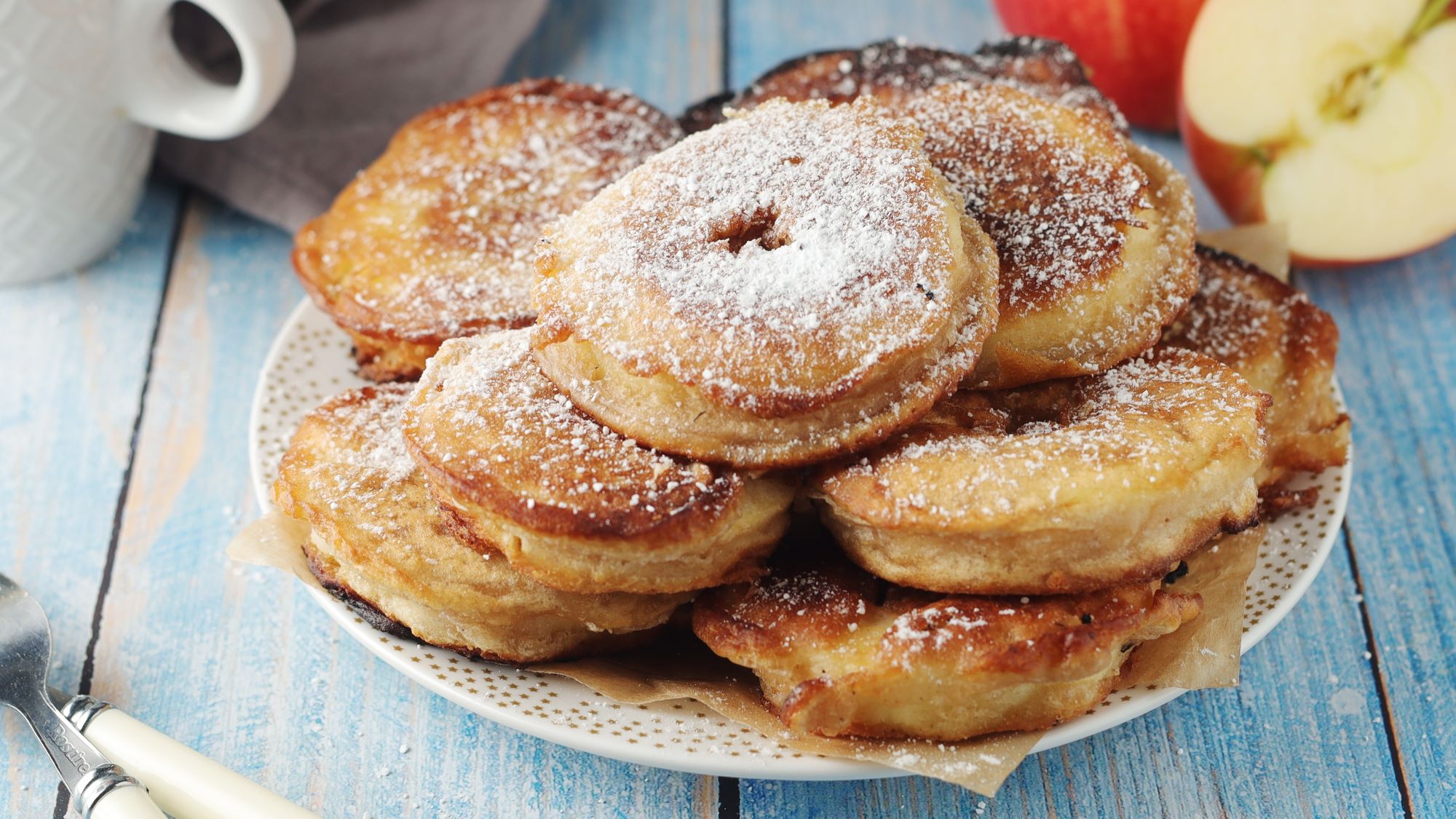Mehrere Apfelplinsen mit Puderzucker bestäubt auf einem gemusterten Teller, daneben ein angeschnittener Apfel.