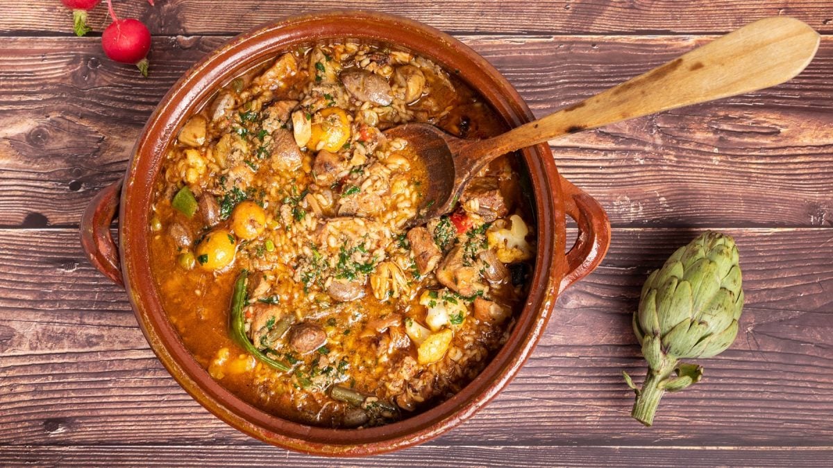 Arroz brut mit zartem Fleisch, buntem Gemüse und Kräutern in Tonform, angerichtet auf gealtertem Holzuntergrund.