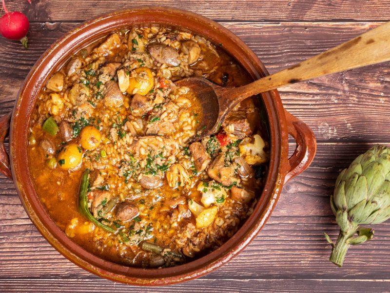 Arroz brut mit zartem Fleisch, buntem Gemüse und Kräutern in Tonform, angerichtet auf gealtertem Holzuntergrund.