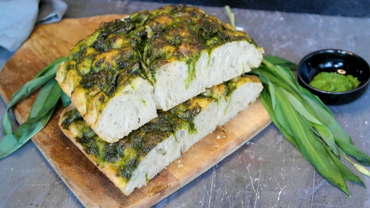 Ein Holzschneidebrett mit der angeschnittenen Bärlauch-Foccacia auf einem grauen Tisch mit frischem Bärlauch und einem schwarzen Schälchen mit grünem Pesto.