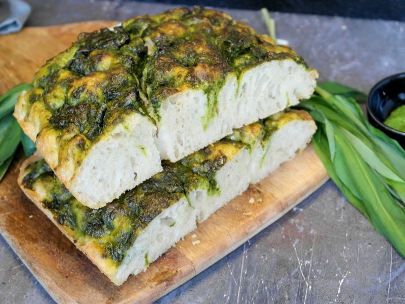 Ein Holzschneidebrett mit der angeschnittenen Bärlauch-Foccacia auf einem grauen Tisch mit frischem Bärlauch und einem schwarzen Schälchen mit grünem Pesto.