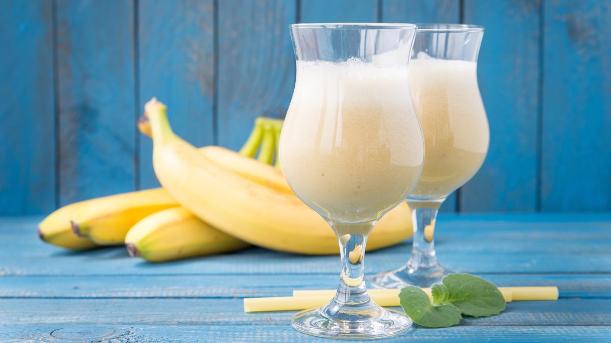 Cremige Sommer-Cocktails mit Banane in bauchigen Gläsern vor gelben Bananen auf strahlend blauem Untergrund serviert.