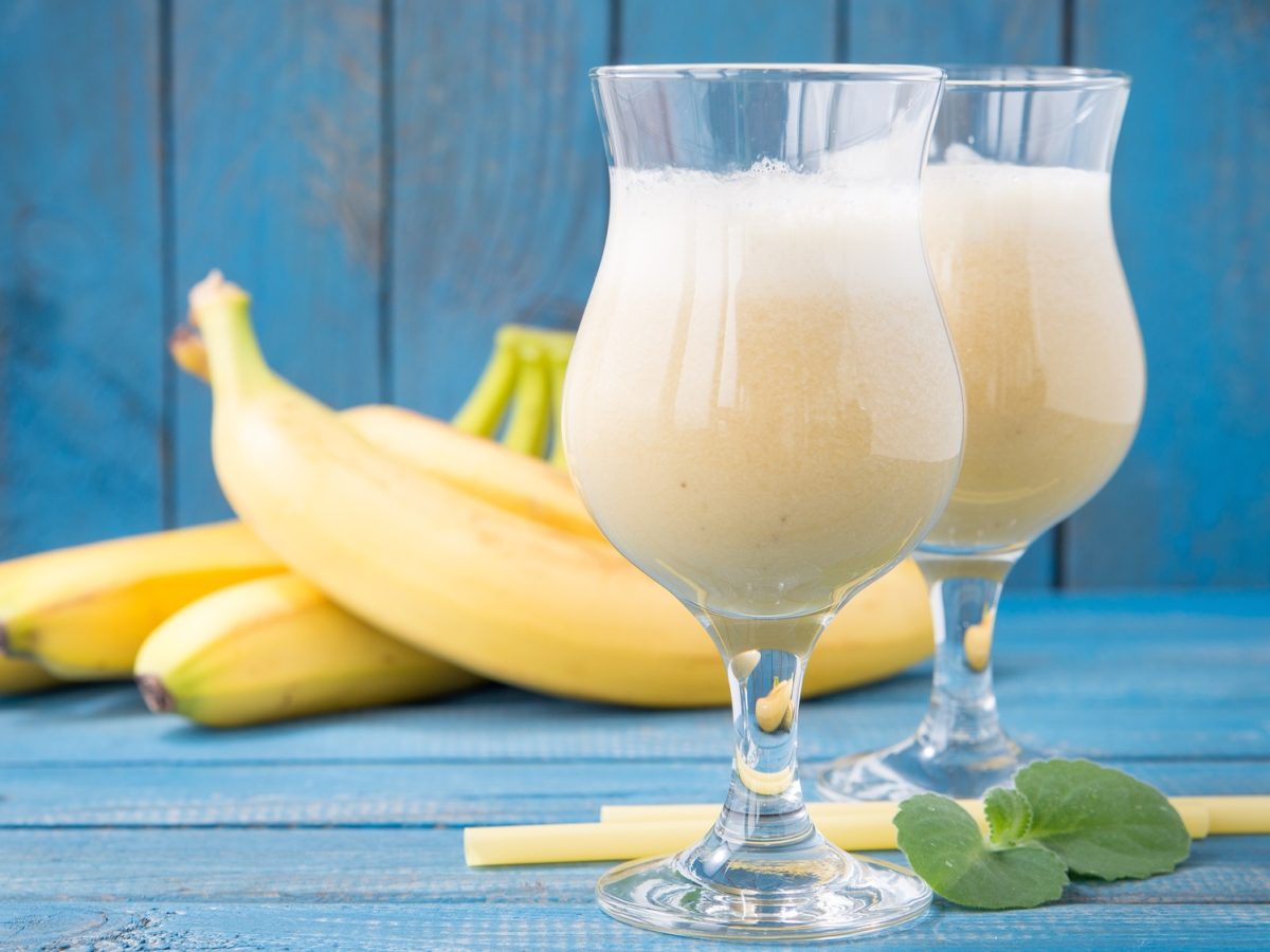 Cremige Sommer-Cocktails mit Banane in bauchigen Gläsern vor gelben Bananen auf strahlend blauem Untergrund serviert.