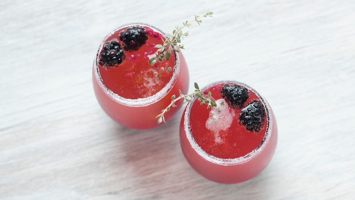 Zwei runde Gläser mit pinkem Brombeer-Thymian-Fizz, dekoriert mit frischen Brombeeren, Thymianzweigen und Zuckerrand.