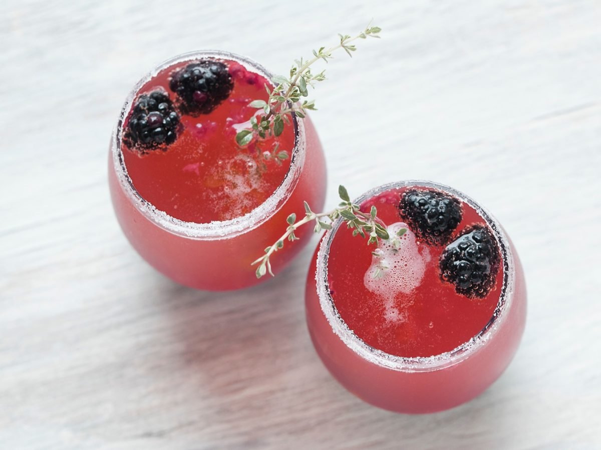 Zwei runde Gläser mit pinkem Brombeer-Thymian-Fizz, dekoriert mit frischen Brombeeren, Thymianzweigen und Zuckerrand.