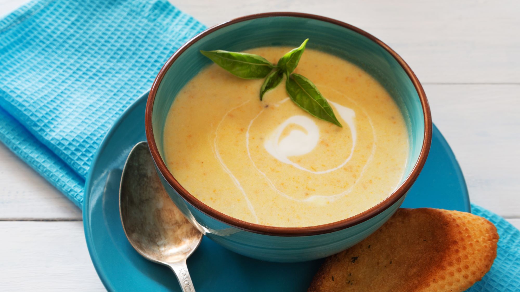 Cremige Buttermilch-Kartoffelsuppe mit Sahnewirbel und Basilikumblättern in türkisfarbener Schale auf blauer Serviette.