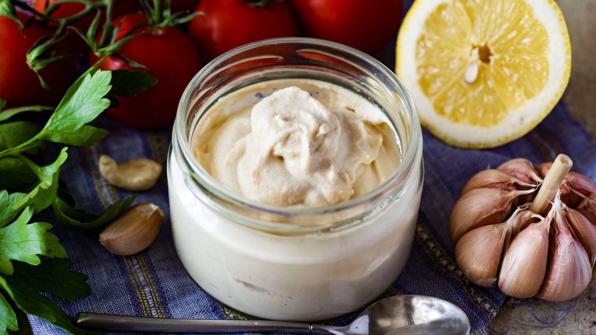 Ein Glas mitd er Mayonnaise auf einem Tisch mit blauer Decke und Salatzutaten im Hintergrund.