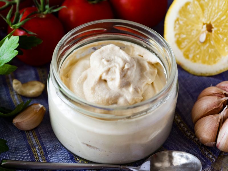 Ein Glas mitd er Mayonnaise auf einem Tisch mit blauer Decke und Salatzutaten im Hintergrund.