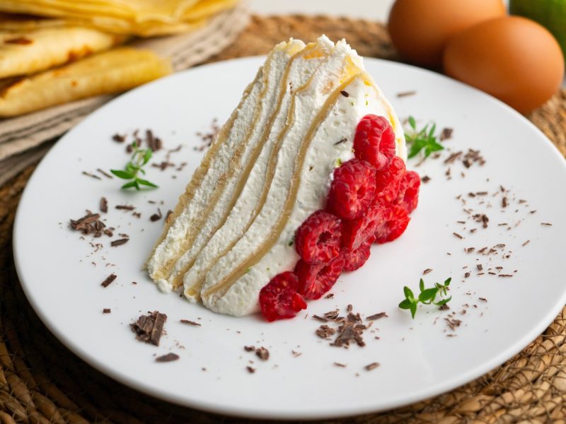 Ein Stück Crêpe-Cake mit Sahneschichten und frischen Himbeeren auf weißem Teller, garniert mit Schokospänen und Kräutern.