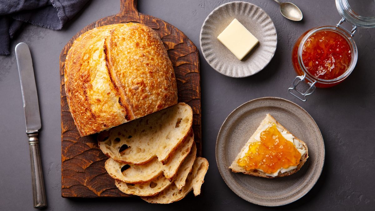 Knuspriges Croissant-Brot mit luftiger Krume liegt auf Holzbrett, serviert mit Butter, Aprikosenmarmelade und grauem Steinteller.