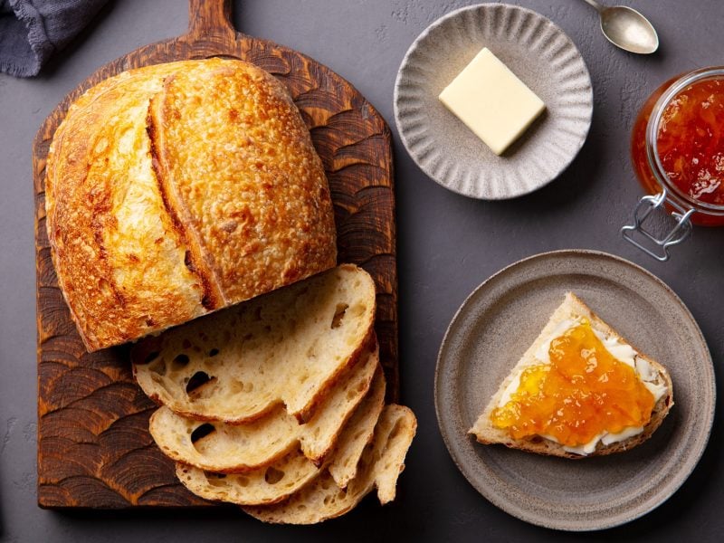 Knuspriges Croissant-Brot mit luftiger Krume liegt auf Holzbrett, serviert mit Butter, Aprikosenmarmelade und grauem Steinteller.