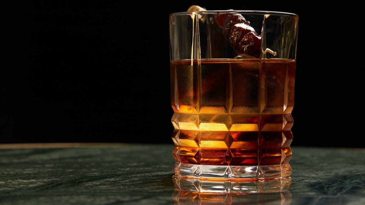 Ein dunkler Cynar Negroni mit Eiswürfeln und Kirsch-Garnitur steht in einem geschliffenen Glas auf grün marmorierter Tischplatte.