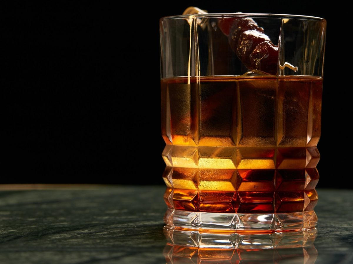 Ein dunkler Cynar Negroni mit Eiswürfeln und Kirsch-Garnitur steht in einem geschliffenen Glas auf grün marmorierter Tischplatte.