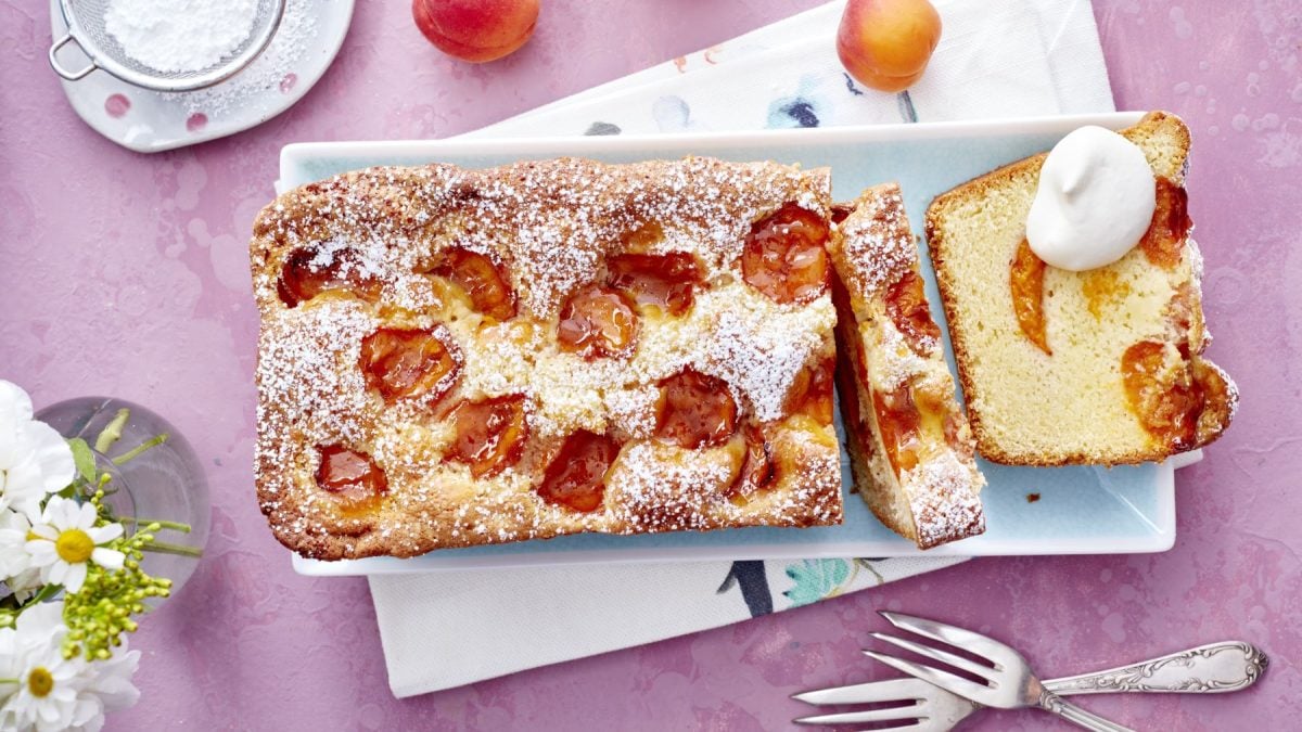 Aprikosen-Limoncello-Kuchen in einer Kastenform, mit frischen Aprikosenstücken und Puderzucker bestäubt, auf einem hellblauen Teller serviert. Daneben eine Kuchenscheibe mit einem Klecks Schlagsahne, auf rosa Untergrund dekoriert mit Gabeln, Blümchen und frischen Aprikosen.