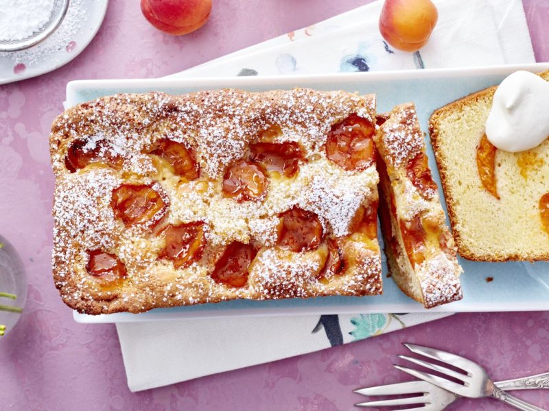 Aprikosen-Limoncello-Kuchen in einer Kastenform, mit frischen Aprikosenstücken und Puderzucker bestäubt, auf einem hellblauen Teller serviert. Daneben eine Kuchenscheibe mit einem Klecks Schlagsahne, auf rosa Untergrund dekoriert mit Gabeln, Blümchen und frischen Aprikosen.