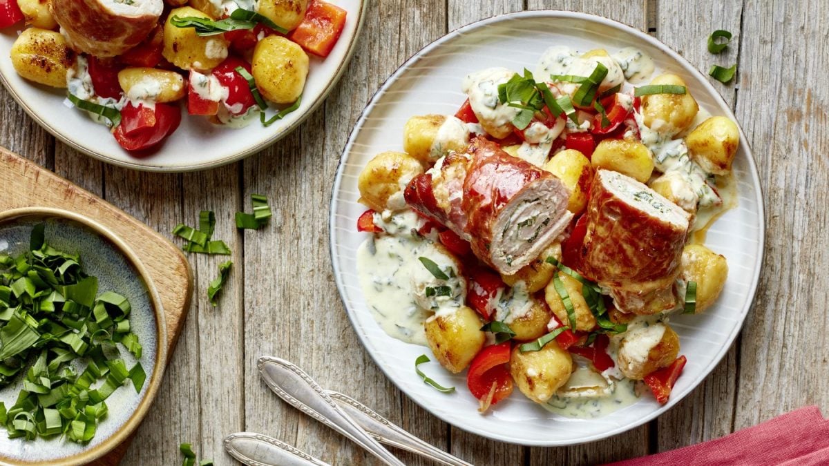 Bärlauch-Schnitzelröllchen auf einem weißen Teller auf hölzernem Hintergrund. Links ein Schälchen mit Bärlauch.