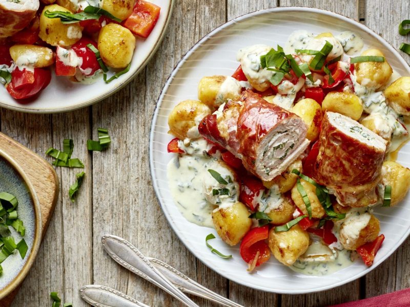 Bärlauch-Schnitzelröllchen auf einem weißen Teller auf hölzernem Hintergrund. Links ein Schälchen mit Bärlauch.