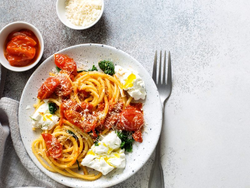 Draufsicht: Ein runder, heller Teller mit Burrata-Pasta mit Tomaten. Daneben liegen eine Gabel und ein Tuch. / zwei kleine runde Schüsseln. Eine mit frischen Tomaten. die andere mit geriebenen Käse.
