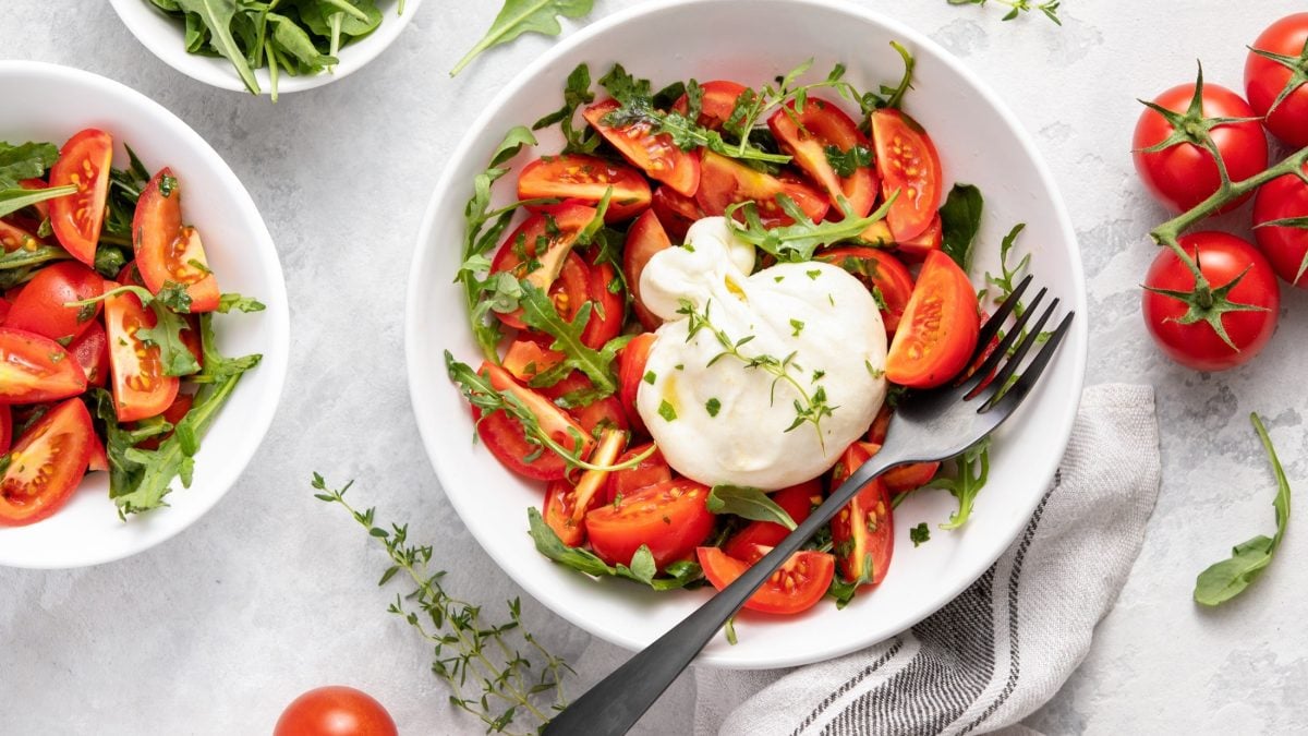 Draufsicht: Zwei Schüsseln mit einem Burrata-Tomaten-Salat und frischem Rucola. Drum herum liegen frische Tomaten.