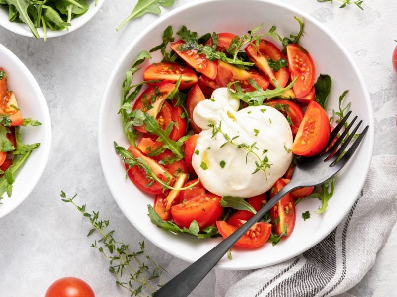 Draufsicht: Zwei Schüsseln mit einem Burrata-Tomaten-Salat und frischem Rucola. Drum herum liegen frische Tomaten.