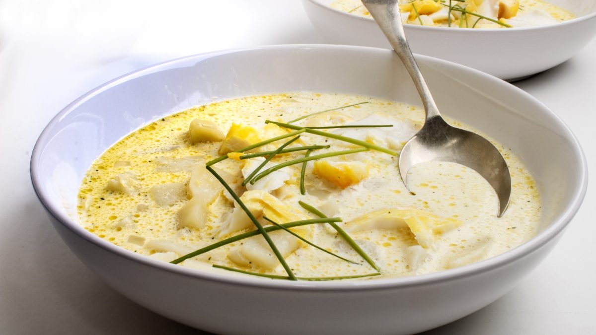 Draufsicht, schräg: Ein runder Teller mit einer Suppe und einem Suppenlöffel auf hellem Untergrund.