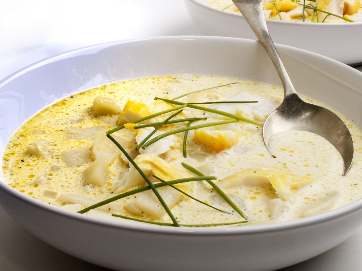 Draufsicht, schräg: Ein runder Teller mit einer Suppe und einem Suppenlöffel auf hellem Untergrund.