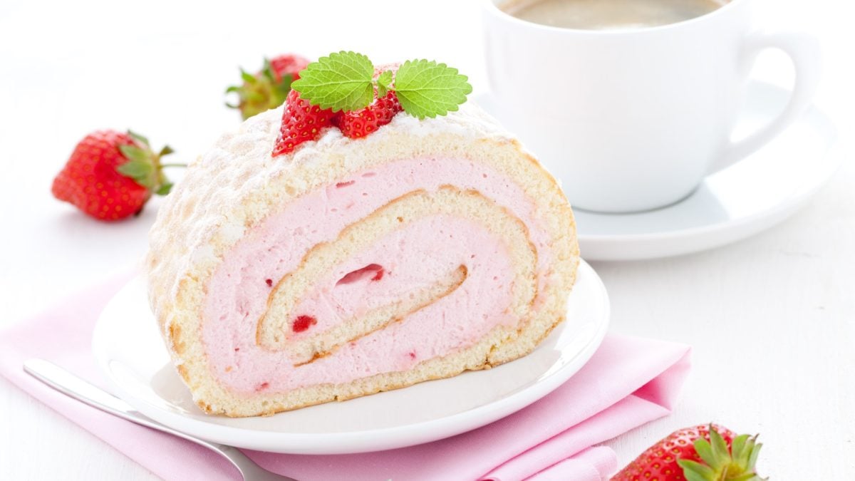 Draufsicht, schräg: Eine cremige Kuchenrolle mit Erdbeeren auf einem hellen Teller. Der liegt auf einem gefaltetem Tuch. Hintergrund: Eine helle Tasse mit einem Getränk auf einem Untersetzer.