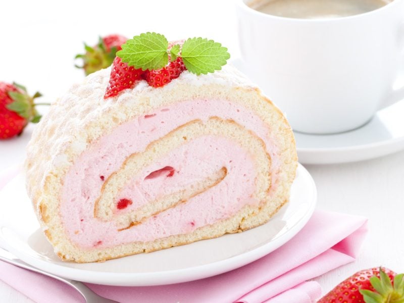 Draufsicht, schräg: Eine cremige Kuchenrolle mit Erdbeeren auf einem hellen Teller. Der liegt auf einem gefaltetem Tuch. Hintergrund: Eine helle Tasse mit einem Getränk auf einem Untersetzer.