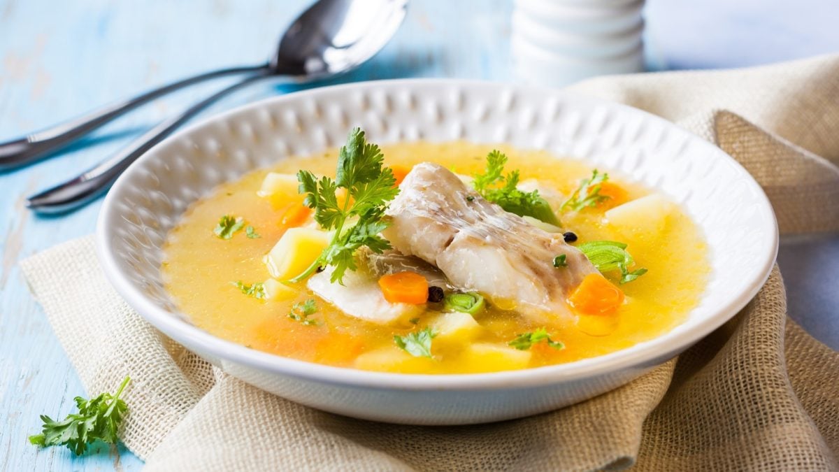 Draufsicht, leicht schräg: Ein runder Teller mit einem Teller Suppe mit Fisch und Gemüse und Besteck.