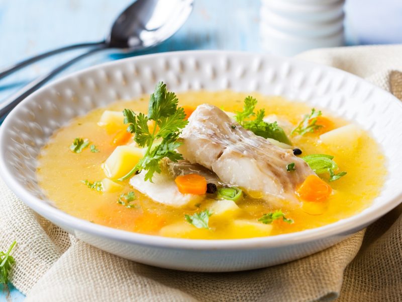 Draufsicht, leicht schräg: Ein runder Teller mit einem Teller Suppe mit Fisch und Gemüse und Besteck.