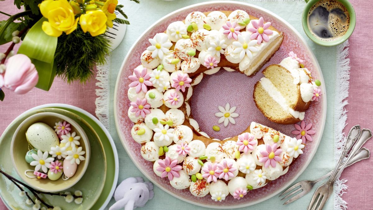 Frühlingskranz auf einem rosa Teller auf einem grünen Geschirrtuch. Rechts Gabeln und links auf dem Foto kleine Teller, Oster-Deko und Blumen. Rechts oben auf dem Foto eine Tasse Kaffee.